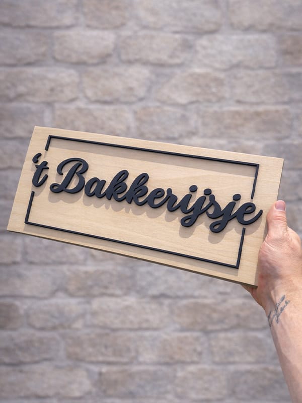 Houten naambord ’t Bakkerijsje met zwarte reliëfletters tegen een lichte bakstenen muur.