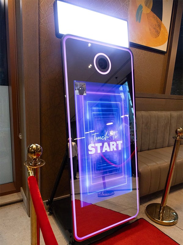 Spiegelfotobooth met neon ‘Touch to Start’-scherm, omringd door rode koorden in een indoor setting.