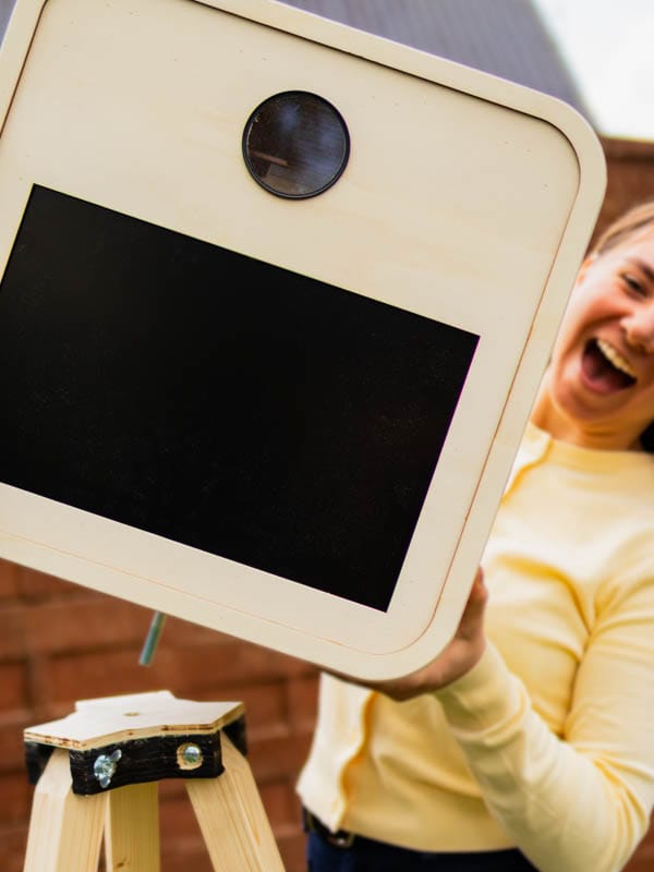 Persoon naast een houten fotobooth op statief tijdens een outdoor event.