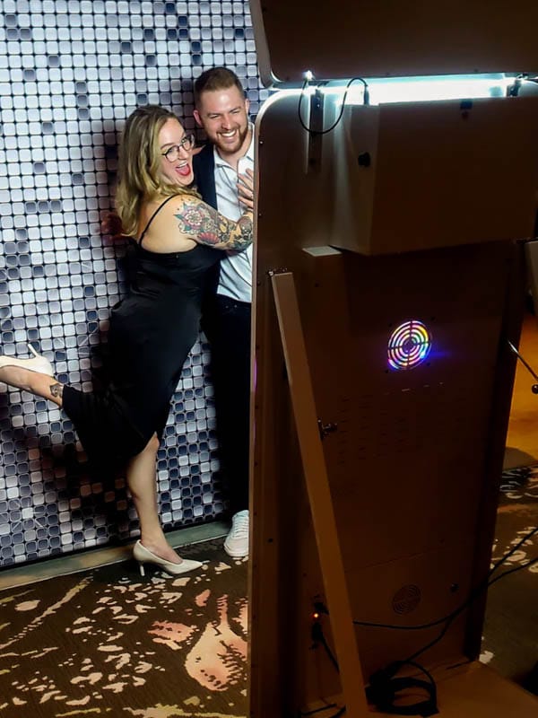 Twee personen poseren voor een fotobooth met kleurrijke verlichting en een reflecterende backdrop.
